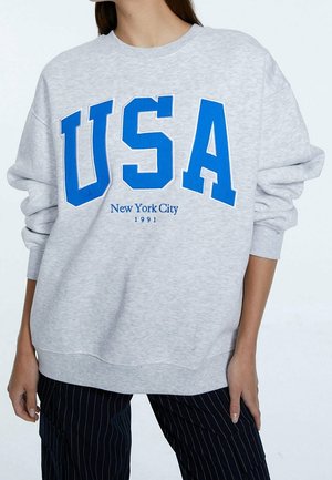 Personne portant un sweat-shirt gris clair avec de grandes lettres bleues "USA" et un texte plus petit "New York City 1991", associé à un pantalon foncé à fines rayures.
