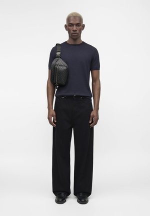 Moški s kratkimi blond lasmi, oblečen v mornarsko modro majico, široke črne hlače, črne čevlje in črno vzorčasto torbico čez ramo.