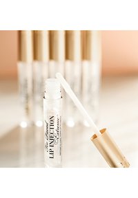 Transparante lipgloss-tube met een gouden dop, voorzien van een witte applicator. Het etiket leest "Too Faced Lip Injection Extreme." De achtergrond bevat andere soortgelijke tubes.