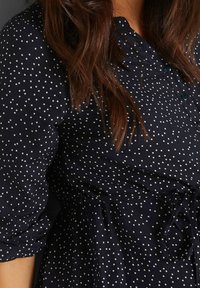 Navyblaues Kleid mit weißen Punkten, ausgestattet mit einem Hemdkragen, elastischem Bund und gerafften Puffärmeln. Der strukturierte Stoff hat einen dezenten Glanz.