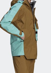 Homme portant une veste à capuche marron et bleu clair avec des poches latérales, debout les mains dans les poches, sur un fond uni.