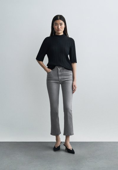 Γυναίκα που στέκεται μπροστά σε απλό τοίχο, ντυμένη με μαύρο πουλόβερ με κοντά μανίκια, γκρι τζιν καμπάνα που φτάνουν ως το ύψος του αστραγάλου και μαύρες μυτερές γόβες.