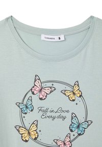 T-shirt di cotone azzurro chiaro con una grafica rotonda che presenta farfalle multicolori e la scritta 'Innamorati ogni giorno' in nero.