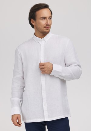 Mężczyzna ubrany w białą, zapinaną na guziki, koszulę z długim rękawem oraz ciemnoniebieskie spodnie, lekko patrzy w prawo, jedną ręką dotyka kołnierza.
