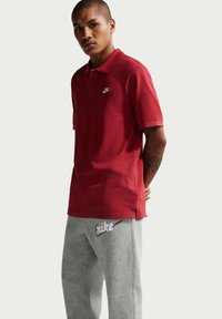 Un bărbat tânăr cu păr scurt, purtând un tricou polo roșu Nike și pantaloni de trening gri Nike, stând cu mâinile la spate.