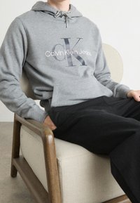 Persona vestida con una sudadera gris de Calvin Klein Jeans y pantalones negros, sentada en una silla de madera acolchada de color beige claro contra una pared lisa.
