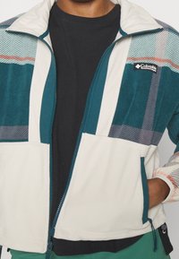 Kurtka polarowa z wzorem w kolorach teal, białym i w kratę, z zamkiem błyskawicznym na przodzie, kieszeniami bocznymi oraz logo Columbia na piersi.