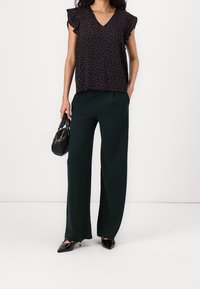 Blusa nera a fiori con maniche arricciate, abbinata a pantaloni a gamba larga di un verde scuro. Accessoriata con una piccola borsa nera e scarpe a punta.