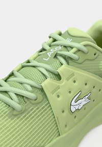 Αθλητικό παπούτσι ανοιχτού πράσινου χρώματος με υφασμάτινο επάνω μέρος με υφή, επίπεδα κορδόνια στο ίδιο χρώμα, λαστιχένιες λεπτομέρειες και λευκή ετικέτα με το λογότυπο Lacoste στη πλευρά.