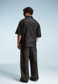 Uomo che indossa una camicia a maniche corte grigio scuro con texture e pantaloni larghi abbinati, scarpe marroni e occhiali da sole neri, in piedi su uno sfondo semplice.