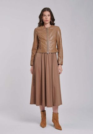Kobieta stojąca w beżowej skórzanej kurtce na zamek, dopasowanej brązowej spódnicy midi i beżowych botkach na jasnym szarym tle.