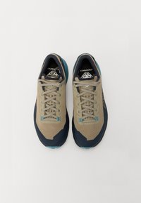 Beige mesh treningssko med marineblå detaljer, som har en teksturert overflate, flate snøringer og såle i gummi med teal-farge.