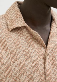 Chemise à motif présentant un design en chevron dans des coloris beige et rouille, avec un col pointu et un tissu texturé.