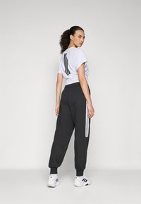 Kvinne med krøllete hår i hestehale iført en hvit t-skjorte med svarte striper, svarte joggebukser med hvite striper, og hvite joggesko.
