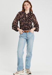 Blouse noire à motifs floraux avec des manches bouffantes, décolleté à nouer, portée avec un jean taille haute bleu clair et des baskets beiges. Une ceinture marron ajoute un contraste.