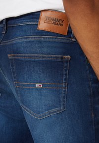 Tmavě modré džíny z denimu s koženou nášivkou označenou "TOMMY JEANS." Mají viditelné švy a malé vyšívané logo na zadní kapse.