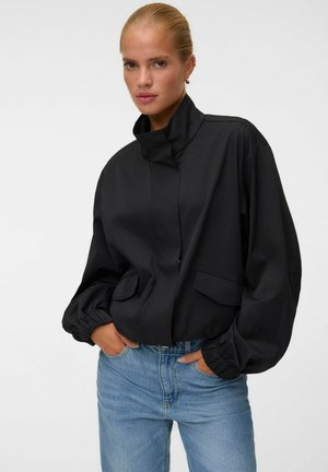 Femme aux cheveux tirés en arrière, portant une veste noire à col haut et un jean bleu, les mains dans les poches de la veste sur fond blanc.