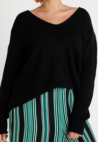Pull noir en tricot avec un col en V et une coupe ample, associé à une jupe rayée verticalement noire et verte. Texture douce et design décontracté.