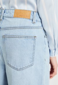 Jeans de denim azul claro com um patch de couro bege rotulado "NOISY MAY". Apresenta dois bolsos traseiros com costura branca e um corte reto.