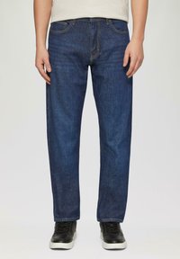 Jeans en denim bleu foncé à coupe droite, avec cinq poches, des coutures jaunes et une légère décoloration. Portés avec des baskets noires.