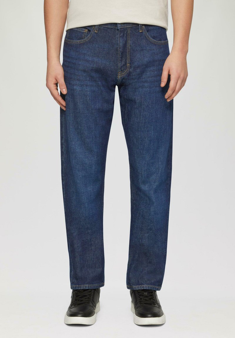 Jeans en denim bleu foncé à coupe droite, avec cinq poches, des coutures jaunes et une légère décoloration. Portés avec des baskets noires.