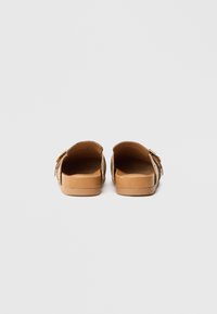 Een paar beige open-achter sandalen met een enkele verstelbare gespsluiting, gezien vanaf de hiel op een witte achtergrond.