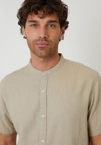 Camisa de manga corta beige de botones en un tejido suave y texturizado, con collar alzado y cinco botones blancos en la parte delantera.