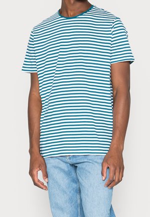 Homme portant une chemise à manches courtes rayée horizontalement turquoise et blanche avec un jean bleu clair, debout devant un fond uni.