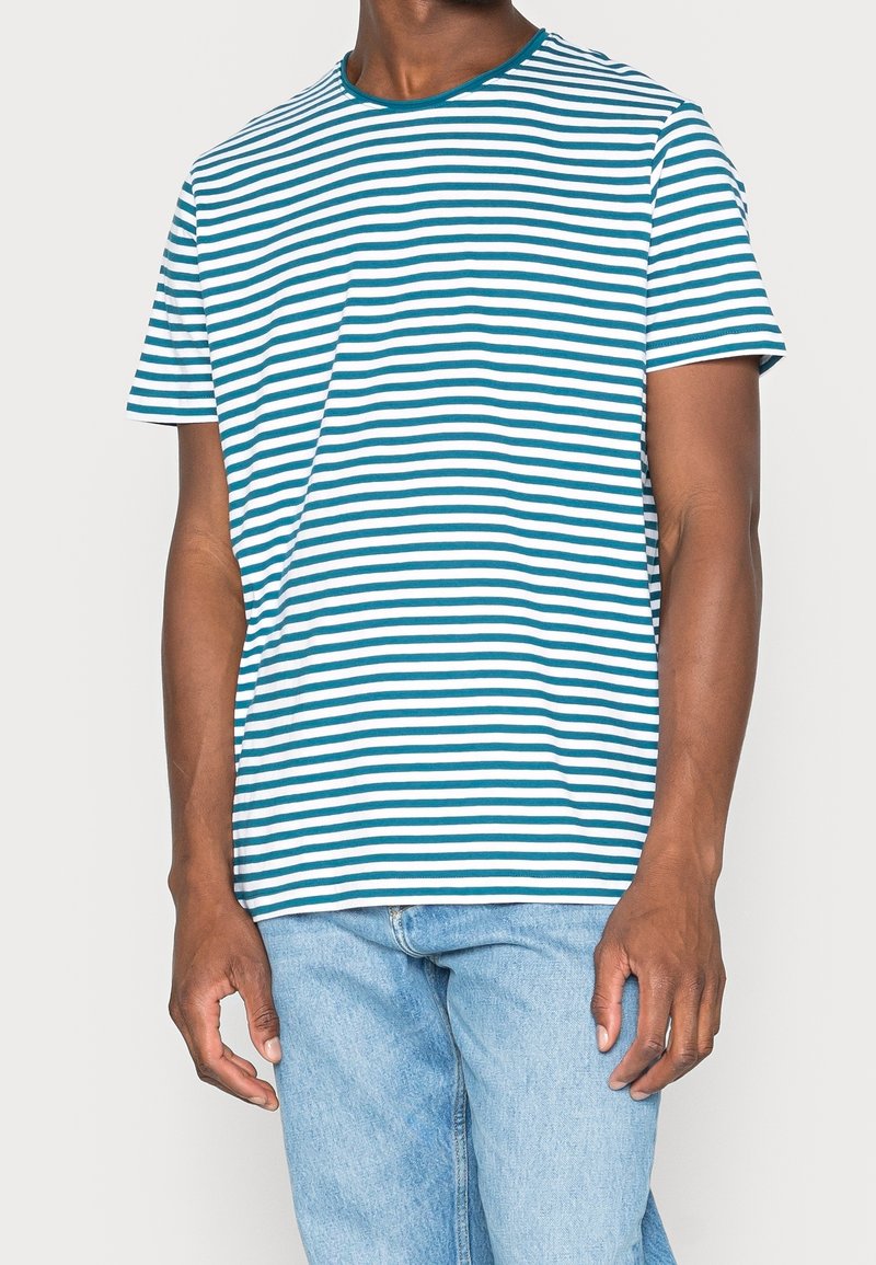 Homme portant une chemise à manches courtes rayée horizontalement turquoise et blanche avec un jean bleu clair, debout devant un fond uni.