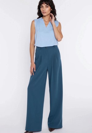 Kobieta o kręconych ciemnych włosach, ubrana w bezrękawową jasnoniebieską bluzkę włożoną w szerokie spodnie o kroju dzwonu w kolorze zielonkawoniebieskim, stojąca na tle białego tła.