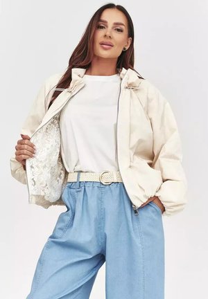Kobieta w jasnobeżowej kurtce z koronkową podszewką, białej koszuli, plecionym pasku i jasnoniebieskich, wysokich spodniach, pozująca z jedną ręką w kieszeni.