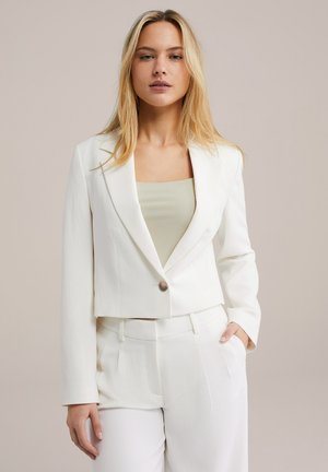 Bílá zkrácená blazer s jedním knoflíkem, kombinovaná se světle zeleným ribbed topem, stylizovaná s odpovídajícími bílými kalhotami.