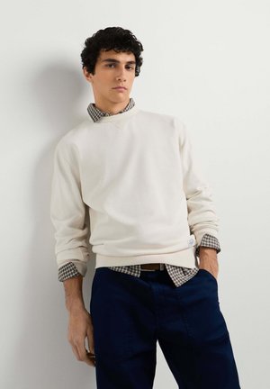 Jonge man die een crèmekleurige trui draagt over een geruit overhemd met marineblauwe broek, staand met één hand in de zak tegen een witte muur.