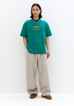 Grøn bomulds t-shirt med trykt design, parret med let beige vidde bukser. Fodtøj i brunt, slip-on stil. Minimalistisk indretning.
