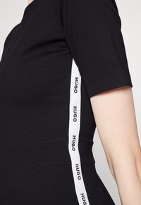 Detailní záběr na černý top s krátkým rukávem, na boku s bílým vertikálním pruhem, na kterém se opakuje černý text "HUGO."