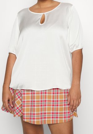 Femme portant un chemisier blanc à manches courtes avec une découpe en forme de larme au niveau du décolleté et une jupe à carreaux beige, rouge et blanc.