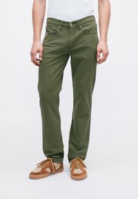 Mann iført olivengrønne straight-leg bukser og brune ruskinds-sneakers med beige detaljer, stående på en ensfarvet hvid baggrund.