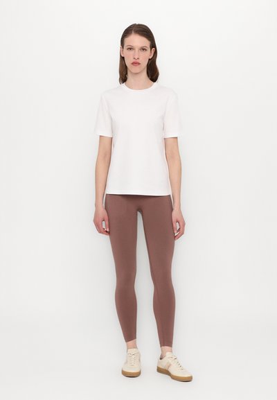 Bela bomba iz bombaža, okroglo izrezana, kratki rokavi, v kombinaciji s svetlo rjavimi oprijetimi leggingi in svetlo bež čevlji na vezanje.