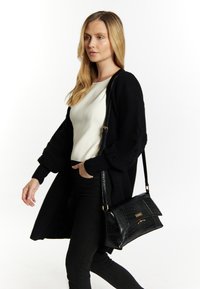 Sort crossbody taske med krokodillemønster, flad design, guld hardware-detaljer, parret med en hvid top og sort cardigan.