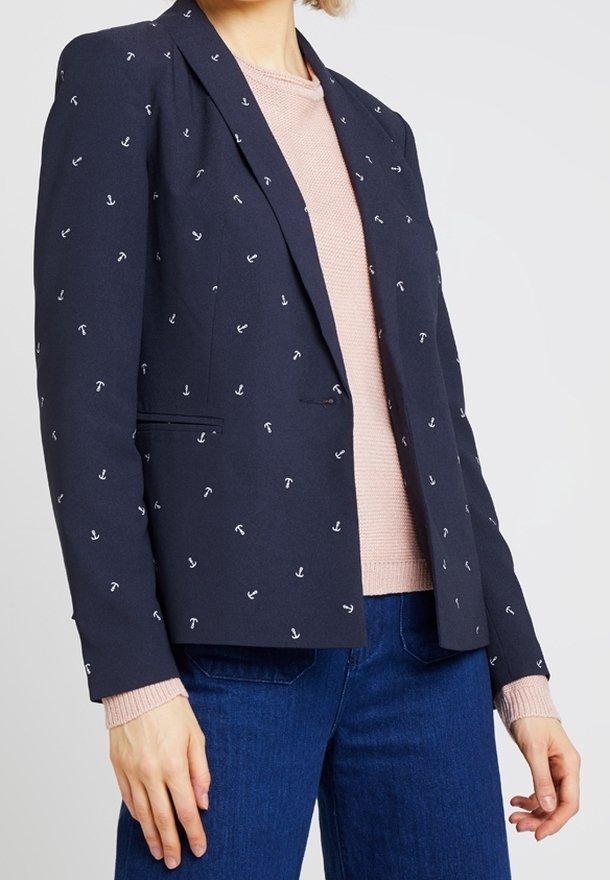 Femme portant un blazer bleu marine avec de petits motifs d’ancre blanche sur un pull rose et un jean bleu, debout avec les bras détendus le long du corps.
