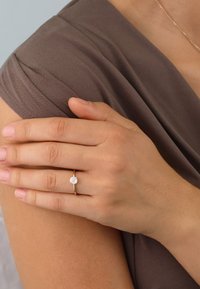 Gold ring with a round, sparkling center stone. Smooth band, set against a soft brown fabric and skin texture. Subtle shine from the metal.