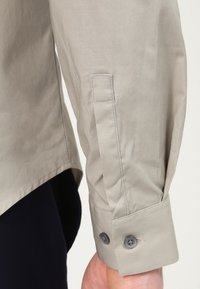 Beige långärmad skjortärm med två knappar och sydd placket, bärs av person med mörka byxor mot en enfärgad bakgrund.