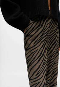 Personne portant un pull noir et un pantalon marron à rayures noires de zèbre, la main glissée dans la poche du pantalon.