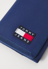 Cartera textil azul con una superficie texturizada. Incluye un parche bordado rectangular con "TOMMY JEANS" en colores rojo, blanco y azul.