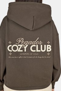 Brauner Kapuzenpullover mit lockerem Schnitt. Mit weißem Drucktext, einschließlich "Pegador COZY CLUB" und dekorativen Motiven darüber und darunter.