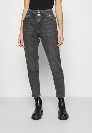 Frau trägt hoch taillierte, ausgewaschene schwarze Jeans mit elastischem Bund und schwarzen Stiefeletten, steht vor einem schlichten hellen Hintergrund.