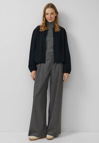 Czarna kurtka bomberka z zamkiem z przodu, szary golf i szerokie szare spodnie. Materiał wydaje się miękki; kurtka posiada elastyczne mankiety i dół. Beżowe buty.