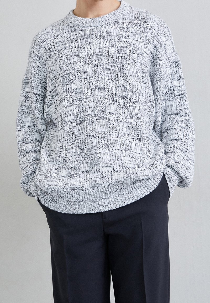 Personne portant un pull gris tricoté texturé avec un motif à carreaux et un pantalon noir, debout les mains dans les poches.