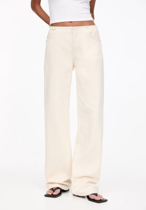 Osoba ubrana w beżowe, szerokie spodnie z wysokim stanem, biały krótki top na ramiączkach oraz czarne sandały na niskim obcasie z kwadratowym noskiem.