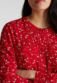 Femme portant un chemisier rouge à boutons avec un petit motif floral blanc, bras croisés, avec de longs cheveux foncés et des lèvres closes visibles.